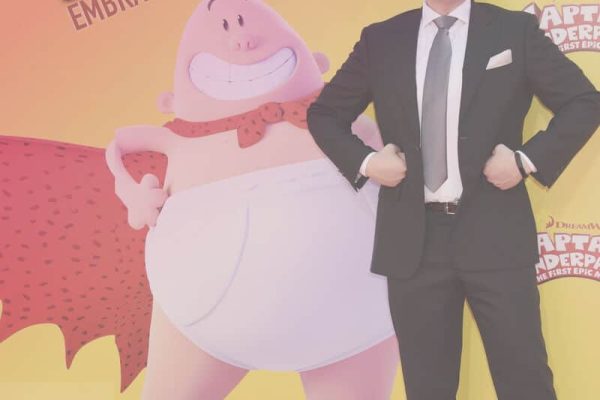 A man in a suit poses confidently on a red carpet in front of a large cartoon superhero image, with text reading "Superdav Embraces Imperfection.